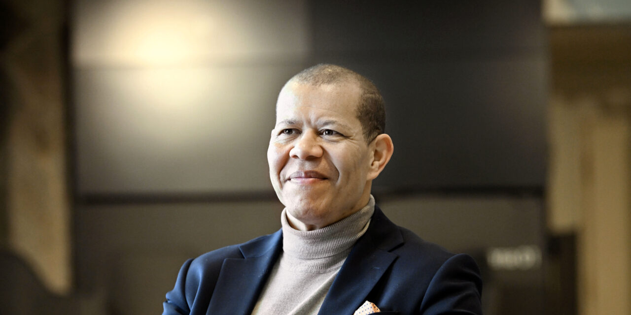 Ari Evwaraye. Henkilö istuu sisätilassa ja hymyilee rauhallisesti. Hänellä on tumma bleiseri ja vaalea poolopaita, ja tausta on pehmeästi valaistu sekä epätarkka.