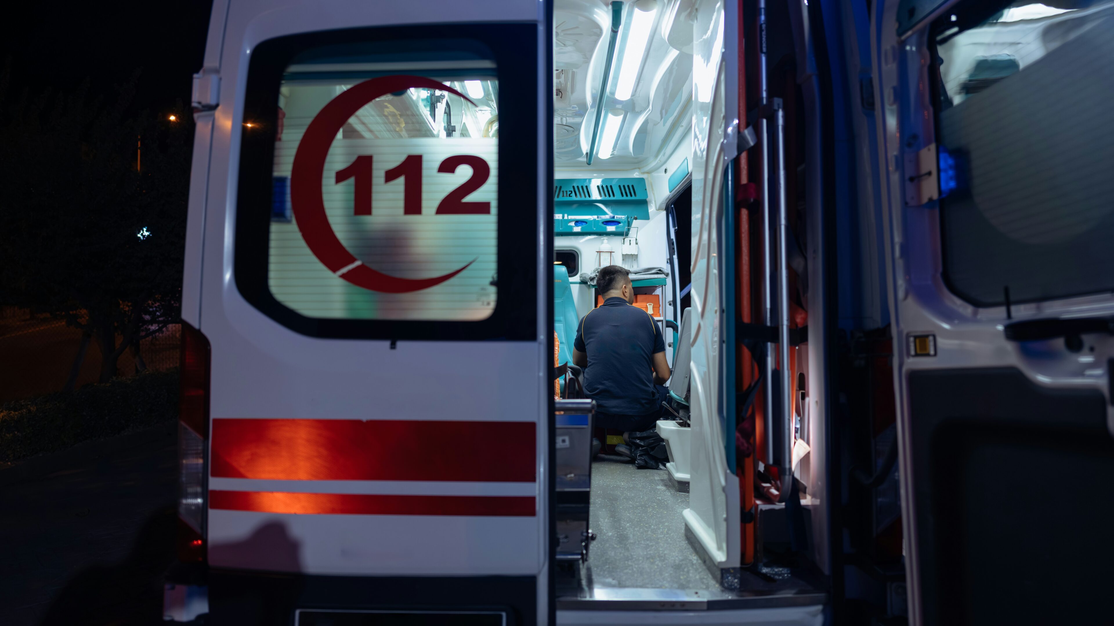 Kuvituskuva: Ambulanssin avoimet takaovet ja valaistu hoitotila, jossa ensihoitaja työskentelee. Takaovessa näkyy 112-tunnus